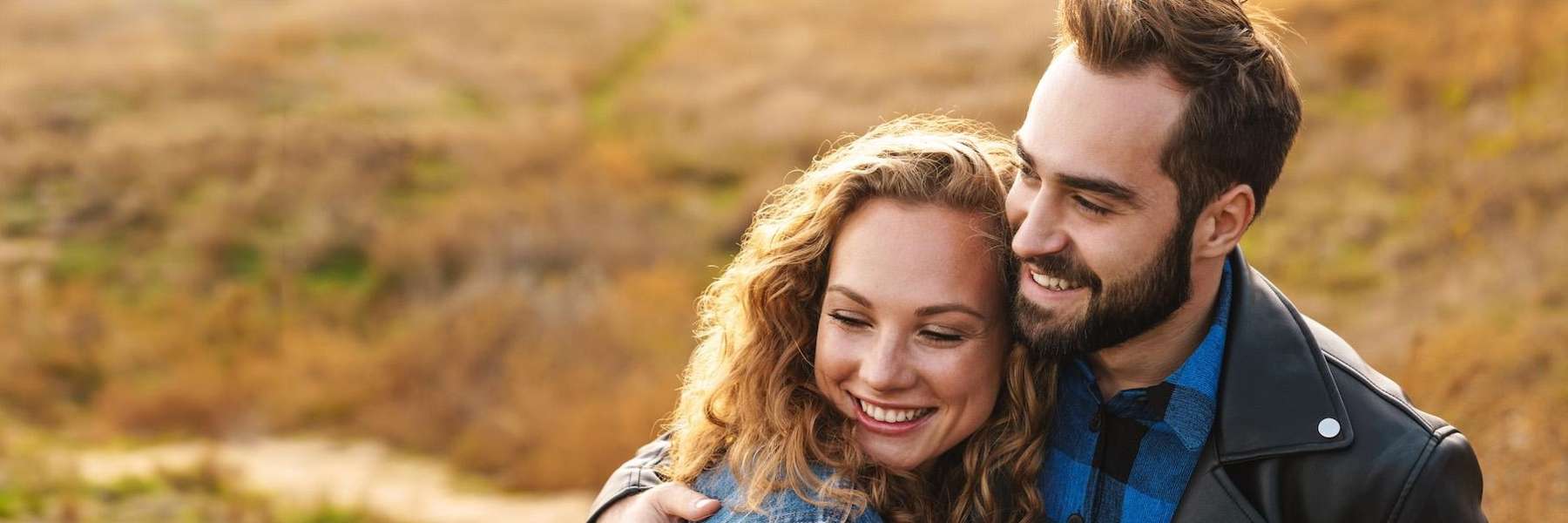 Man and woman embrace in a sunlit field, surrounded by lush greenery and blooming flowers.